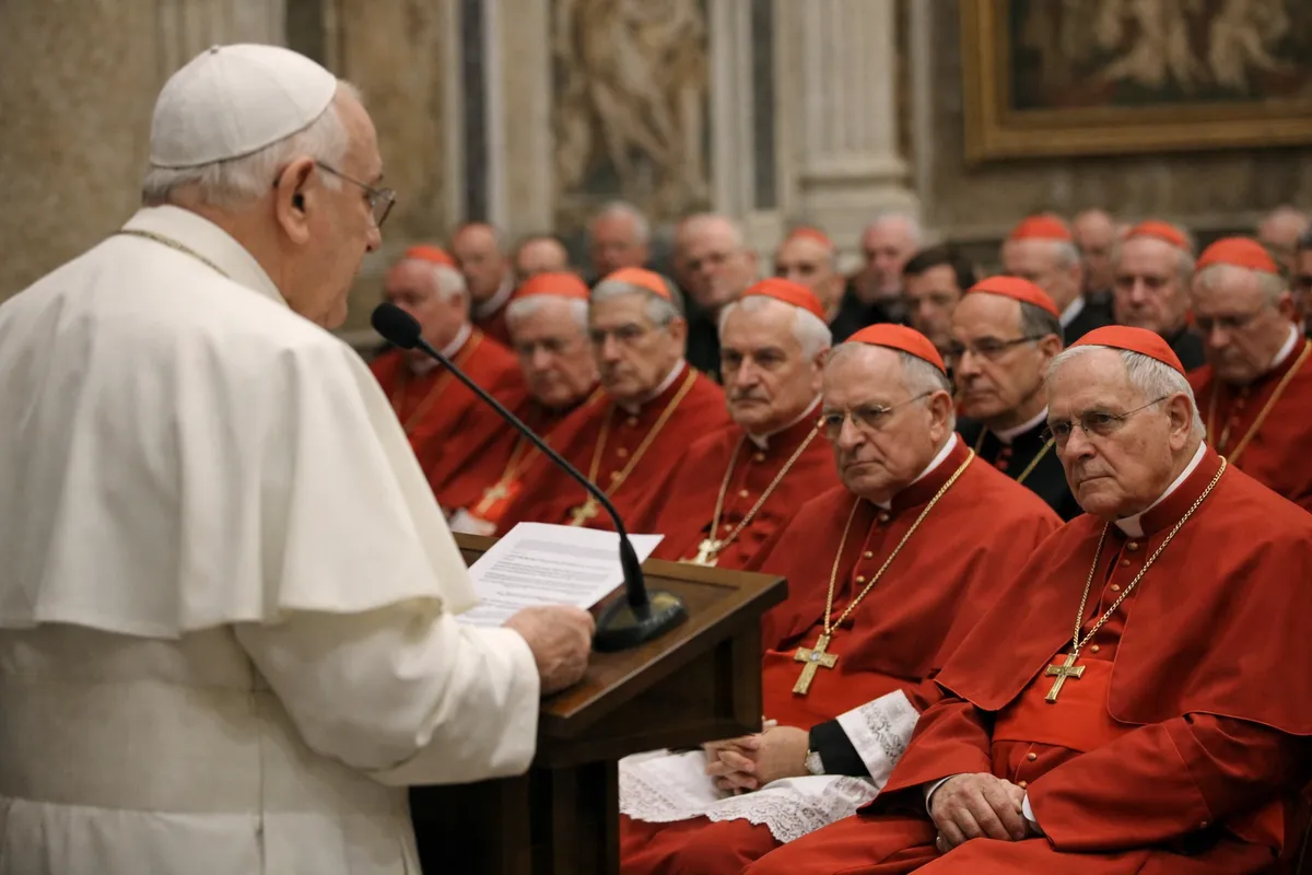 Anziano pontefice in bianco parla al leggio davanti a cardinali in rosso seduti in una sala vaticana