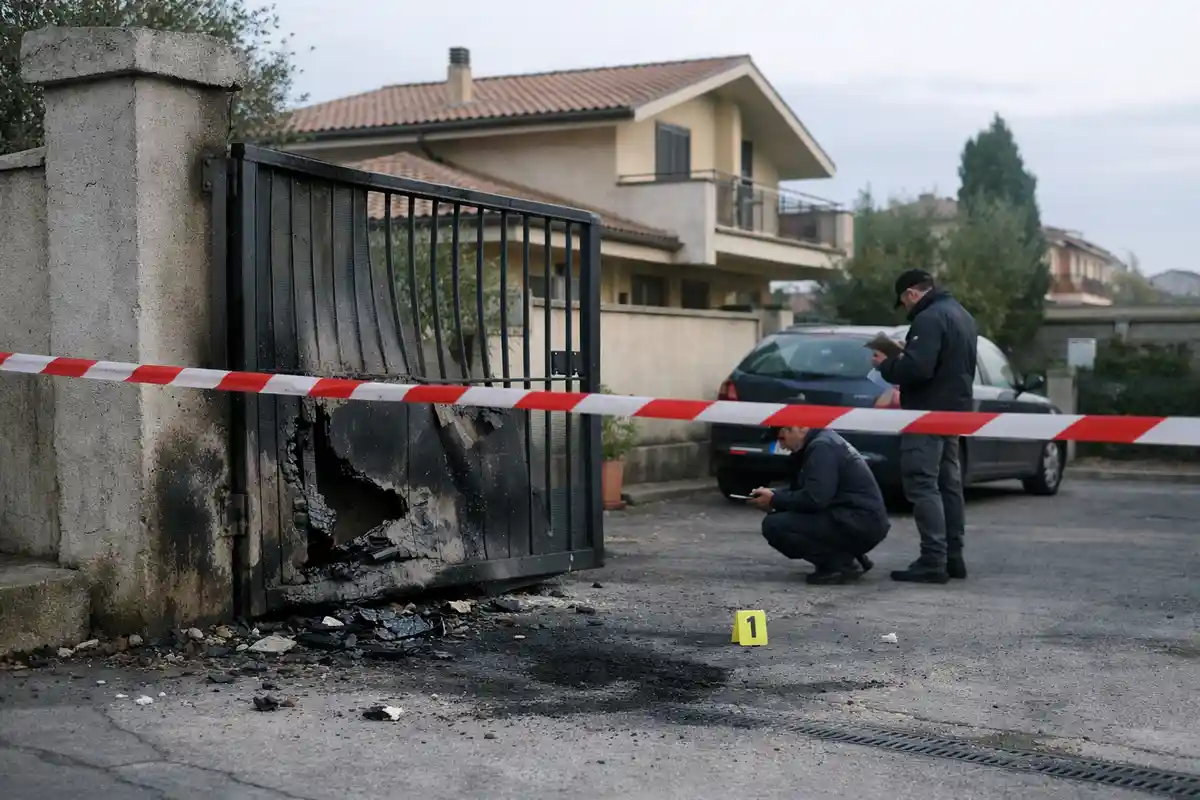 Cancello in ferro bruciato e deformato con nastro della polizia e investigatori al lavoro davanti a una casa